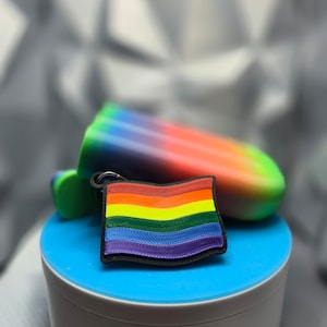 May include: A rainbow-colored pride flag keychain with a textured surface. The keychain is on a blue and white round container.
