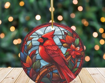 May include: A round Christmas ornament featuring a vibrant red cardinal bird perched on a branch, set against a stained-glass style background with blue, orange, and brown hues. The ornament has a gold string for hanging.