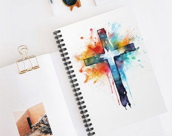 Op de afbeelding: Spiraalgebonden notitieboek met een aquarel kruisontwerp. Het kruis is weergegeven in tinten blauw, groen en zwart, met een levendige splash van rode, oranje, gele en turquoise verf. Het notitieboek is open en toont blanco pagina's.