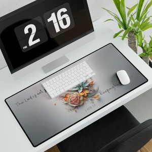 May include: A gray desk mat with a floral design and the text "The Lord is not keeping his promise" and "2 Peter 3:9". A white keyboard and mouse are on the mat. A computer monitor and plant are in the background.