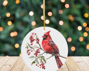 May include: A round, white ceramic ornament featuring a vibrant red cardinal perched on a branch with red berries and green leaves. The ornament hangs from a gold string, perfect for holiday decor.
