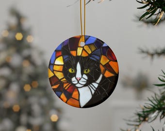 May include: A round ceramic ornament featuring a black and white cat with green eyes. The cat's face is surrounded by a stained glass design with colorful geometric shapes in shades of blue, yellow, orange, and red.