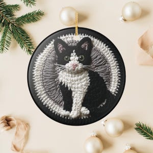 以下が含まれることがあります： 緑色の目とピンクの鼻を持つ白黒の猫が描かれた丸いクリスマスオーナメント。 猫は、白とグレーのステッチのボーダーが付いた黒い背景に配置されています。 金色の吊り下げ紐が含まれています。 お祝いの装飾。
