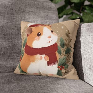 May include: A square cushion with a cartoon guinea pig wearing a red scarf and hat. The guinea pig is set against a beige background with a wreath of green leaves and red berries. The cushion is on a grey sofa.