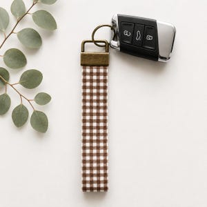 May include: A fabric wristlet keychain featuring a brown and white gingham check pattern. The strap is attached to a rectangular antique brass-toned metal clasp and a circular split ring, which holds a black and silver automotive key fob.