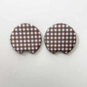 May include: Two brown and white checkered car coasters. The coasters are round with a small notch on one side. The pattern is a classic gingham design. The coasters are designed to fit in a car's cup holder.