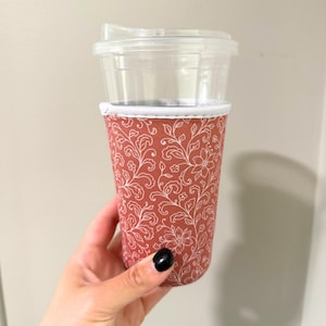 May include: A reusable coffee cup with a clear plastic lid and a terracotta-colored sleeve. The sleeve features a white floral pattern. The cup is held by a person with a black painted fingernail.