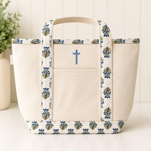 May include: A cream-colored canvas tote bag featuring two handles and a central exterior pocket. The bag is accented with a blue and green floral block print pattern along the base, handles, and top trim. A small blue cross is embroidered on the front.
