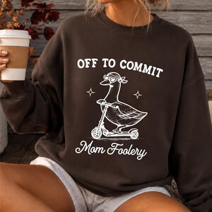 May include: A dark brown crewneck sweatshirt featuring a white graphic of a duck wearing heart-shaped sunglasses and a bandana while riding a kick scooter. The text above reads OFF TO COMMIT and the text below reads Mom Foolery in white font.