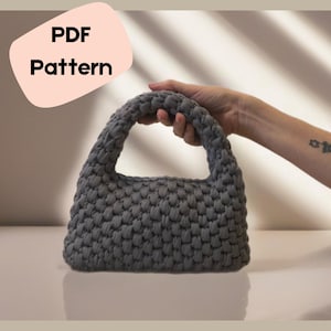 Könnte beinhalten: Eine graue gehäkelte Handtasche mit einer strukturierten Oberfläche. Die Tasche hat einen einzigen Henkel und ist aus einem dicken Garn gefertigt.