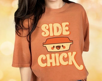 May include: A burnt orange t-shirt with the text "Side Chick" in a retro font. The text is accompanied by a cartoon image of a casserole dish with a smiling face.
