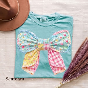 May include: A seafoam green t-shirt featuring a large, central bow applique made from various fabric patches. The bow includes floral patterns in blue, yellow, and pink, alongside a pink and white gingham check section, all with ruffled edges.