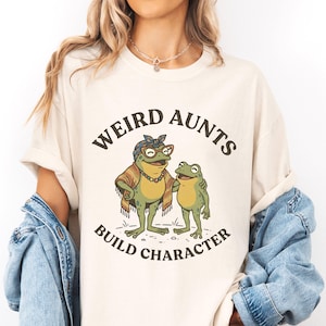 May include: Cream-colored t-shirt with the text "WEIRD AUNTS BUILD CHARACTER." The graphic features two cartoon frogs, one wearing glasses and a shawl, the other with its arm around the first. The shirt is a casual, unisex style.