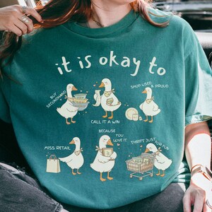 Pu&ograve; includere: T-shirt color verde acqua con la frase "it is okay to" e illustrazioni di oche. Le oche sono raffigurate con oggetti legati allo shopping e testo come "Buy Secondhand" e "Thrift Just for Fun".