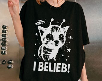 May include: Black t-shirt featuring a white graphic of a cat with antennae and UFOs, surrounded by stars. The text "I BELIEB!" is printed below the cat. The shirt is a casual, unisex style.