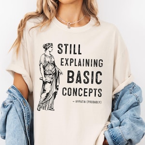 May include: Cream-colored t-shirt with the text "STILL EXPLAINING BASIC CONCEPTS - HYPATIA (PROBABLY)" in black. A black and white illustration of a classical figure is on the left side of the shirt.