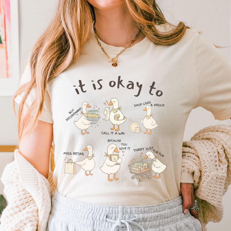 Pu&ograve; includere: T-shirt color crema con la frase "it is okay to" sopra una serie di anatre dei cartoni animati. Ogni anatra illustra un aspetto diverso del thrift shopping, come l'acquisto di seconda mano e lo shopping di usato. La maglietta ha uno scollo tondo.