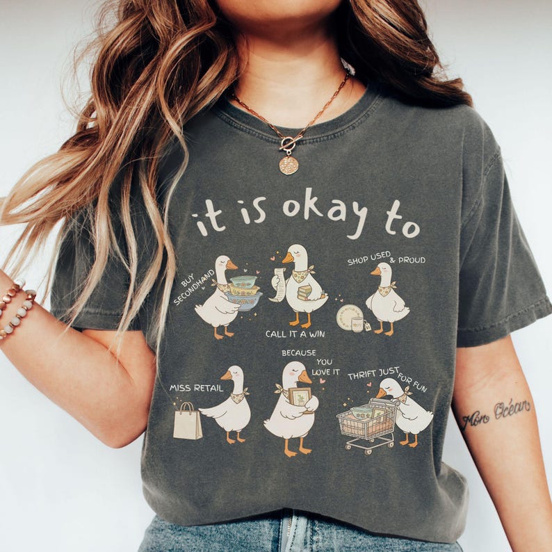 Pu&ograve; includere: T-shirt grigio scuro con la frase "it is okay to" sopra illustrazioni di oche. Le oche sono raffigurate con oggetti legati allo shopping e testo come "Buy Secondhand" e "Thrift Just for Fun."