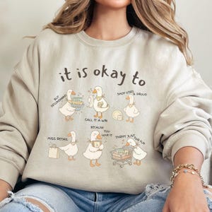 Pu&ograve; includere: Felpa beige con la scritta "it is okay to" e illustrazioni di oche. Le oche sono raffigurate con borse della spesa, piatti e carrelli, con frasi come "Buy Secondhand" e "Thrift Just for Fun".