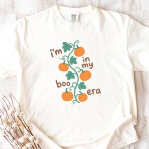 May include: Cream-colored t-shirt with the text "I'm in my boo era" in brown and green. The design features a vine with orange pumpkins and green leaves, perfect for fall or Halloween.