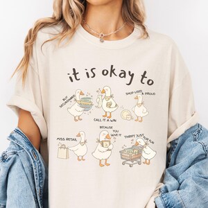 Pu&ograve; includere: T-shirt color crema con la scritta "it is okay to" sopra illustrazioni di oche dei cartoni animati. Le oche sono raffigurate con vari oggetti e frasi, tra cui "Buy Secondhand" e "Thrift Just for Fun".