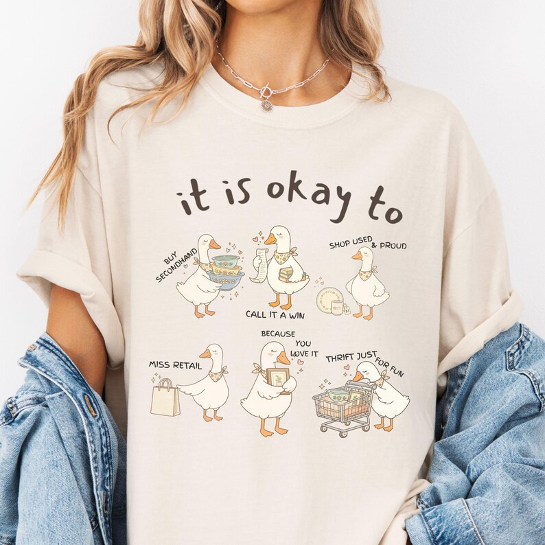 Pu&ograve; includere: T-shirt color crema con la scritta "it is okay to" sopra illustrazioni di oche dei cartoni animati. Le oche sono raffigurate con vari oggetti e frasi, tra cui "Buy Secondhand" e "Thrift Just for Fun".