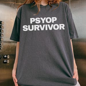 May include: Dark gray t-shirt with the white text "PSYOP SURVIVOR". The shirt is a relaxed fit and appears to be made of a soft material. The person is wearing striped pants.