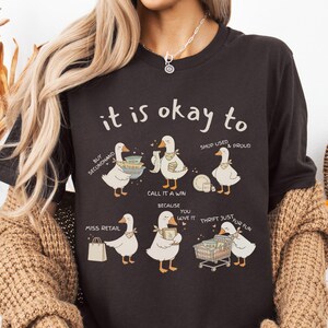 Pu&ograve; includere: T-shirt marrone scuro con la scritta "it is okay to" sopra illustrazioni di oche. Le oche sono raffigurate con vari oggetti, tra cui piatti, un carrello della spesa e una borsa della spesa. La maglietta promuove il riciclaggio e l'acquisto di seconda mano.