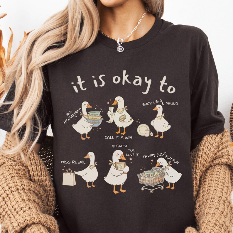 Pu&ograve; includere: T-shirt marrone scuro con la scritta "it is okay to" sopra illustrazioni di oche. Le oche sono raffigurate con vari oggetti, tra cui piatti, un carrello della spesa e una borsa della spesa. La maglietta promuove il riciclaggio e l'acquisto di seconda mano.