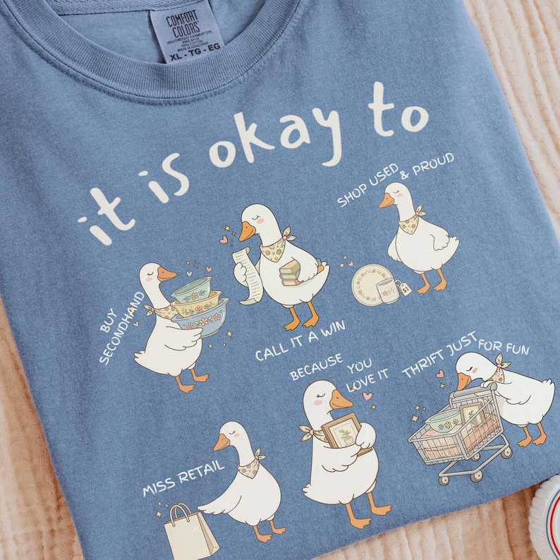 Pu&ograve; includere: Una maglietta blu Comfort Colors con la frase "it is okay to" e illustrazioni di oche. Le oche sono raffigurate con carrelli della spesa, che tengono oggetti e le scritte "Buy Secondhand", "Shop Used & Proud", "Call it a Win", "Miss Retail" e "Thrift Just for Fun".