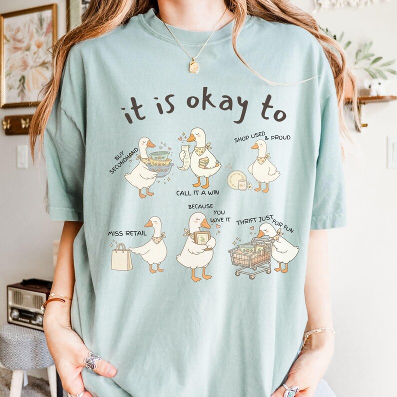 Pu&ograve; includere: Maglietta azzurra con la frase "it is okay to" e illustrazioni di oche. Le oche hanno fumetti con frasi come "Compra usato", "Fai acquisti usati e orgogliosi" e "Risparmia solo per divertimento".
