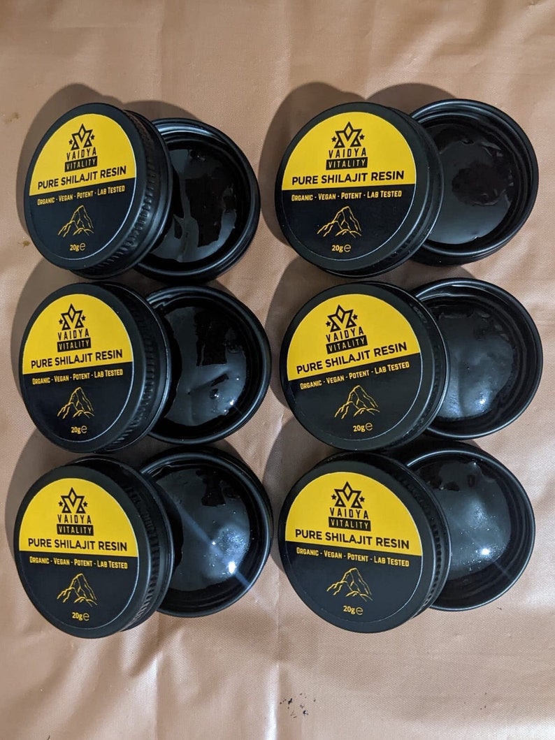 May include: Six circular black containers of Vaidya Vitality Pure Shilajit Resin, each with a yellow lid featuring the brand logo, text, and a mountain graphic. Each lid is labeled 20g, and the open containers reveal a dark, viscous substance.
