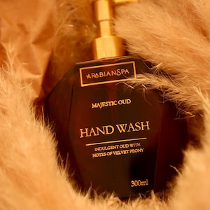 Puede incluir: Una botella de jabón de manos de vidrio marrón oscuro con una bomba dorada. La botella tiene una etiqueta dorada con el texto "Arabian Spa", "Majestic Oud", "Hand Wash", "Indulgent Oud with Notes of Velvet Peony" y "300ml".