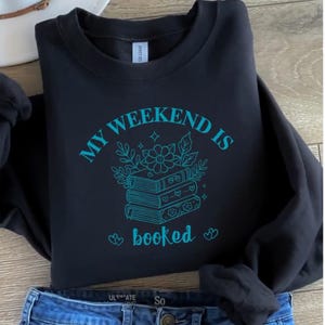 May include: Black sweatshirt with the text "MY WEEKEND IS booked" in teal. The graphic features a stack of books with floral and leaf accents. The sweatshirt is folded, with a pair of blue jeans visible at the bottom.