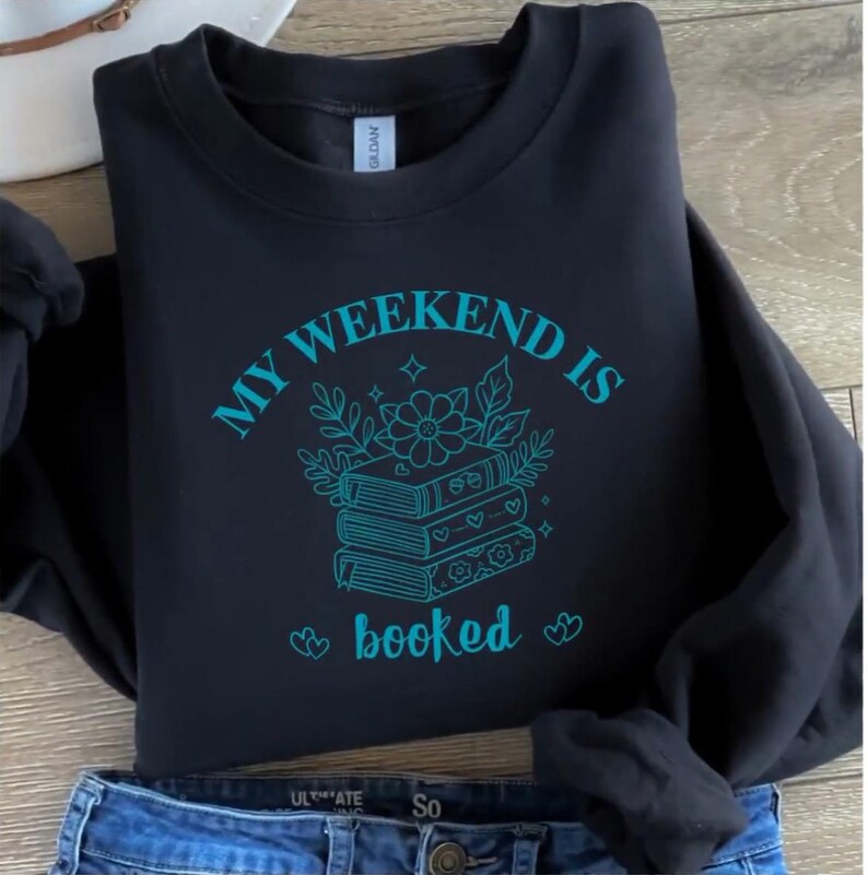 May include: Black sweatshirt with the text "MY WEEKEND IS booked" in teal. The graphic features a stack of books with floral and leaf accents. The sweatshirt is folded, with a pair of blue jeans visible at the bottom.