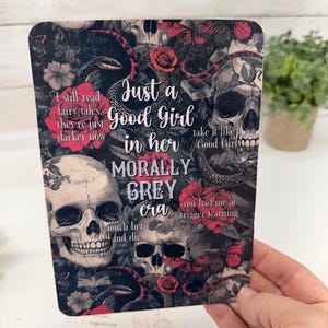 May include: A rectangular card featuring a dark, gothic-inspired print with skulls, red roses, butterflies, and snakes. The text reads: "I still read fairy tales, they're just darker now", "Just a Good Girl in her MORALLY GREY era", "take it like a Good Girl", "you had me at trigger warning", and "touch her and die".
