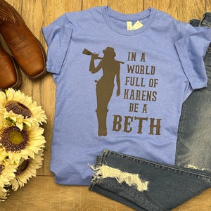 May include: A light blue t-shirt with a brown silhouette of a woman holding a rifle. The text "In a world full of Karens be a Beth" is printed on the shirt.