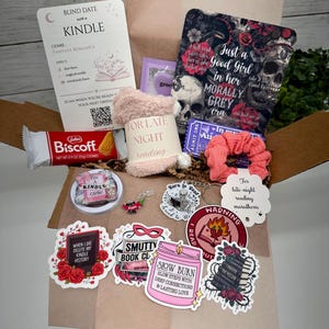 May include: A gift box containing a Blind Date with a Kindle card, a Biscoff cookie, fuzzy socks, a pink scrunchie, a purple ticket, and various stickers featuring skulls, books, and text like "Smutty Book Club" and "Slow Burn" on a brown paper base.