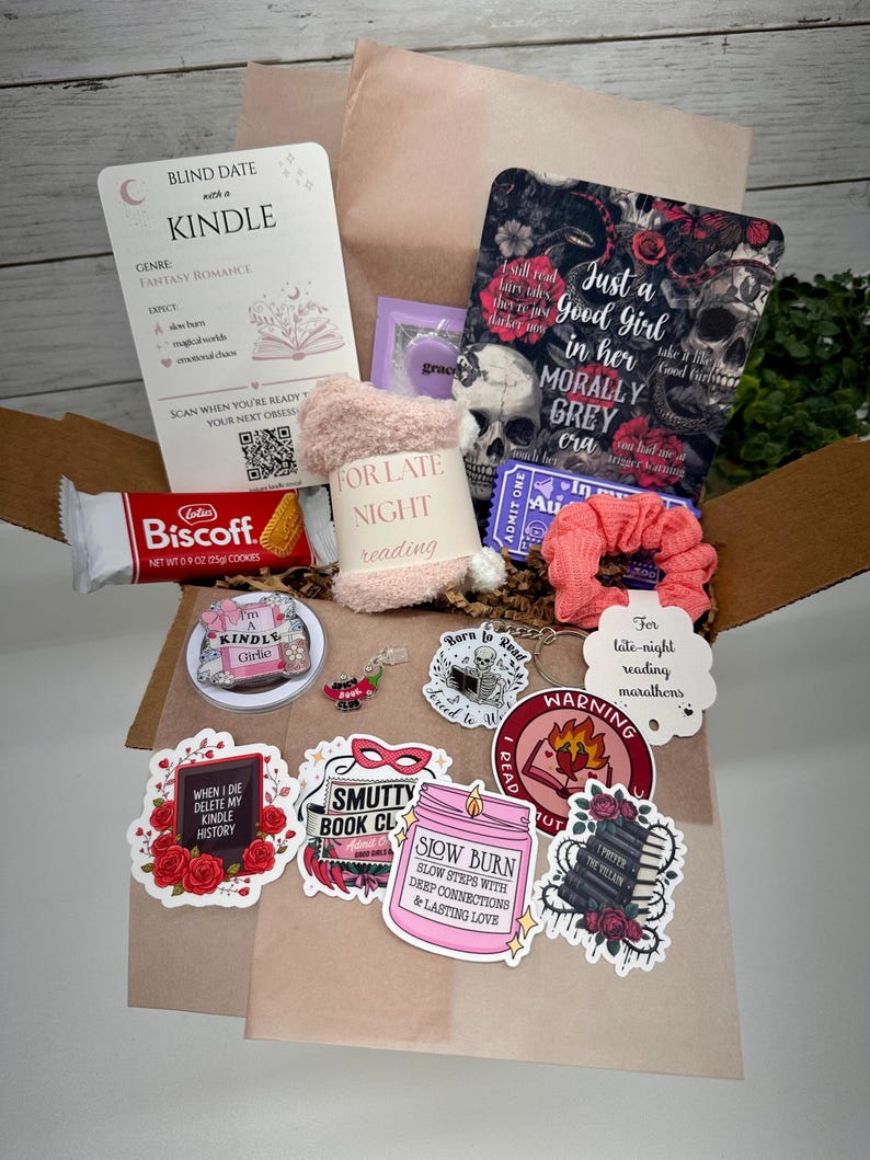 May include: A gift box containing a Blind Date with a Kindle card, a Biscoff cookie, fuzzy socks, a pink scrunchie, a purple ticket, and various stickers featuring skulls, books, and text like "Smutty Book Club" and "Slow Burn" on a brown paper base.