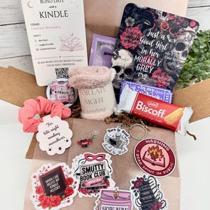 May include: A cardboard box containing a Blind Date with a Kindle card, a skull-themed book cover, a pink fuzzy mug sleeve, a Biscoff cookie, a pink scrunchie, a metal keychain, and various stickers with text including Smutty Book Club and Warning.