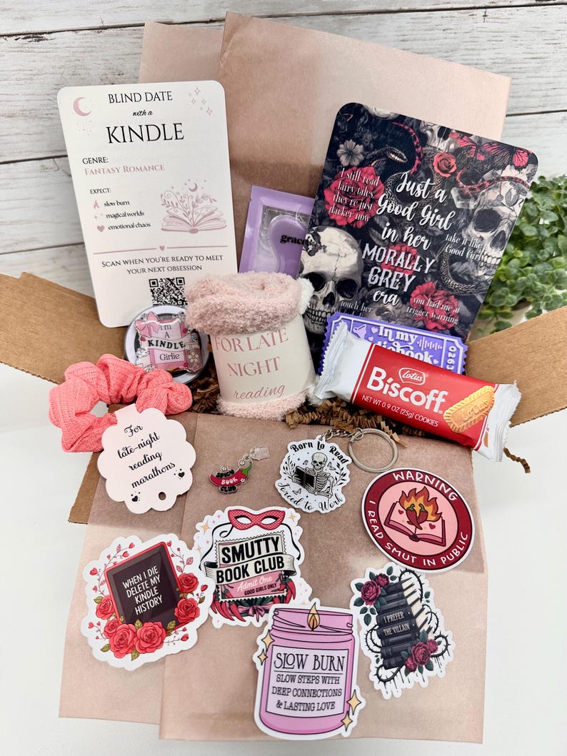 May include: A cardboard box containing a Blind Date with a Kindle card, a skull-themed book cover, a pink fuzzy mug sleeve, a Biscoff cookie, a pink scrunchie, a metal keychain, and various stickers with text including Smutty Book Club and Warning.
