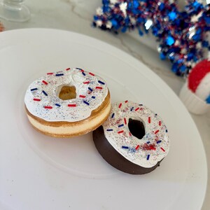 May include: Two donuts on a white plate, decorated with white frosting and red, white, and blue sprinkles. One donut has a light brown base, and the other has a dark brown base. The donuts are themed for a patriotic holiday.