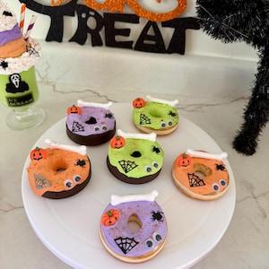 May include: A white plate holds six Halloween-themed donuts with colorful frosting. Each donut is decorated with a pumpkin, spiderweb, and spider. The donuts have orange, green, and purple frosting with chocolate bases. A black cat decoration is in the background.