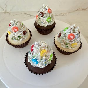 May include: Four cupcakes with white frosting, colorful sprinkles, and M&M candies. The cupcakes have a chocolate base and are displayed on a white plate. The frosting is swirled and decorated with various colored candies.