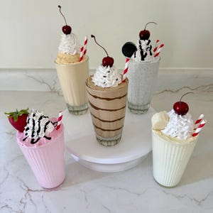 May include: Five decorative milkshake glasses, each with a different flavor and topping. The milkshakes are topped with whipped cream, cherries, and other garnishes like strawberries, bananas, and cookies. Each glass has a red and white striped straw.