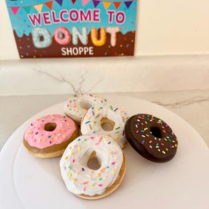 May include: A collection of colorful donuts with various toppings, including sprinkles and chocolate frosting, are displayed on a white surface. A sign above reads "WELCOME TO DONUT SHOPPE" in a playful font, suggesting a bakery or dessert theme.