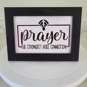 May include: A framed print with the word "prayer" in cursive, and the text "The Strongest Wifi Connection" in a black frame. The image includes a wifi symbol with a cross on top. The frame is sitting on a white surface.