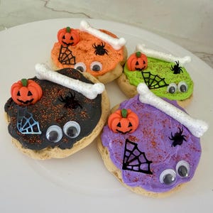 May include: A plate of Halloween-themed cookies. Each cookie is decorated with colored frosting, a pumpkin, a bone, a spider, and googly eyes. The frosting colors include orange, green, black, and purple.