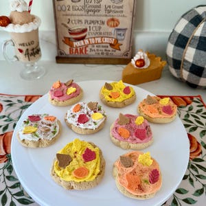 May include: A white serving plate holds eight miniature fall-themed cookies, each topped with colorful frosting and leaf-shaped decorations. A decorative drink and a slice of pie are also visible. A sign in the background reads "Hello Autumn".