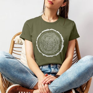 May include: A woman wearing a green t-shirt with a white tree ring design. She is sitting in a wicker chair with her legs crossed and wearing blue jeans.